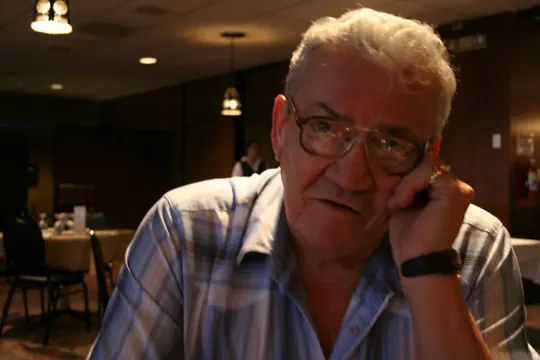 An elderly man in a plaid shirt rests his chin on his hand, seated in a dimly lit restaurant with pendant lights and blurred figures in the.