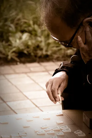 An elderly person contemplates domino pieces on a tiled surface outdoors.