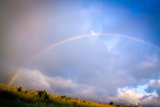 A vibrant rainbow arches over a golden field under a partly cloudy sky.