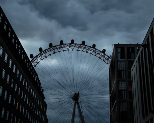 Giant Ferris wheel rises between dark buildings under brooding storm clouds.
