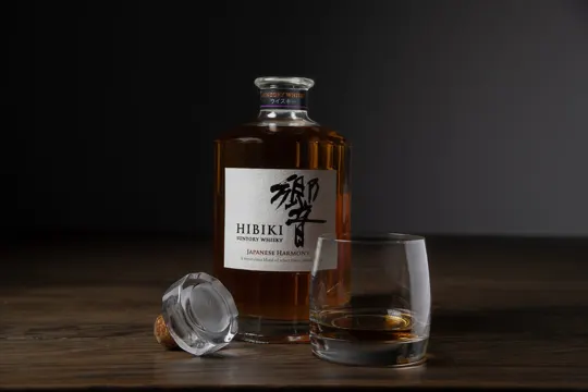 A bottle of Hibiki Japanese Harmony whisky, glass with amber liquid, and a cork, set on a dark wooden surface with soft, directional.