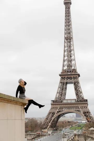 A woman sits on a ledge, gazing skyward against the Eiffel Tower's lattice under overcast light.