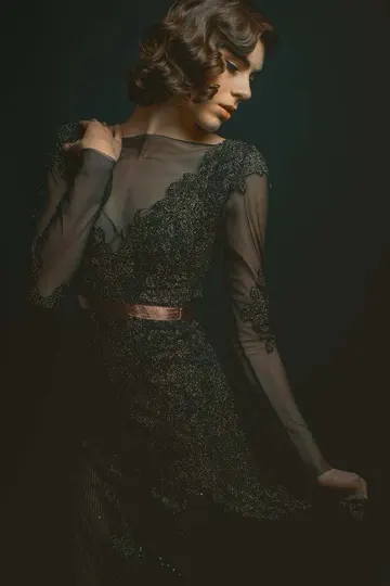A woman in a black lace dress with sheer sleeves and a copper belt stands against a dark background, illuminated by soft, directional light.