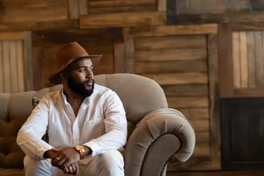 Man in white shirt and brown hat sits on beige sofa, wooden backdrop.