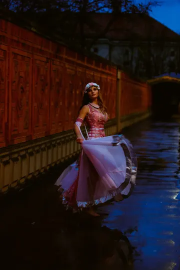 A dancer in a pink gown stands on a wet stone path beside a red wall, illuminated by twilight.