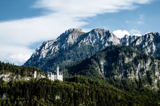 A castle perches on a forested hillside beneath jagged, sunlit mountains.