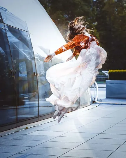 A dancer in a sequined top and flowing white skirt leaps mid-air near a reflective glass building, sunlight illuminating her form.