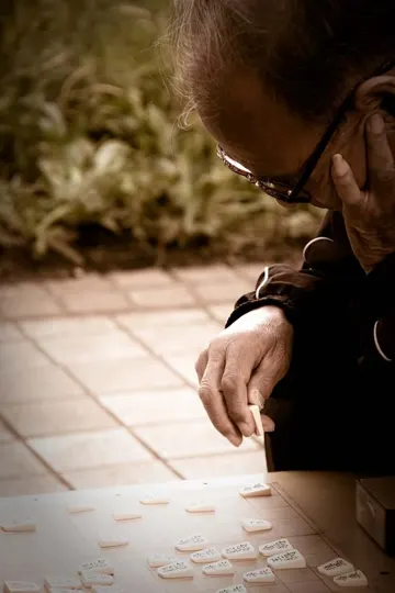 An elderly person contemplates domino tiles outdoors under soft, natural light.