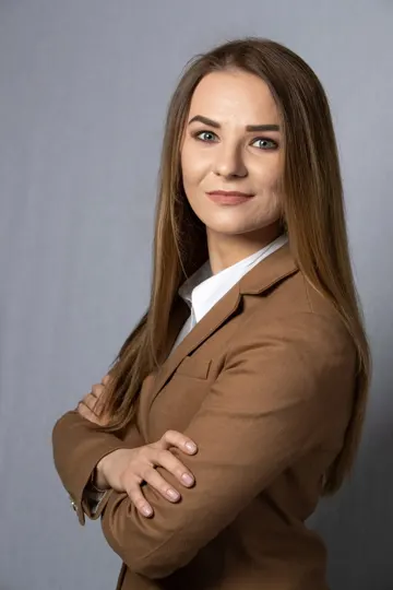 A woman in a brown blazer stands against a neutral gray backdrop.