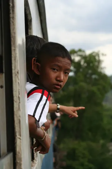 A young boy leans out of a train window, pointing towards a distant, green landscape under a cloudy sky.