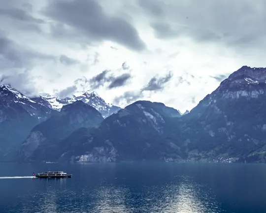 A ferry glides across a lake, framed by snow-capped mountains under a moody, overcast sky.