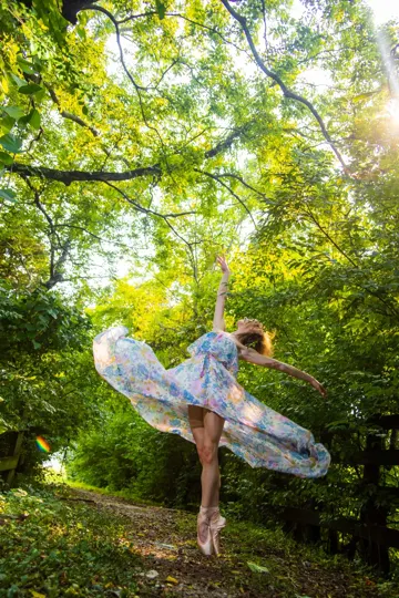 Dancer in floral dress pirouettes on forest path, sunlight filters through green canopy.