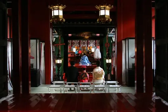 Two individuals kneel before a Shinto shrine, illuminated by warm lanterns.
