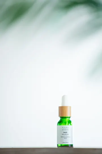 A green glass bottle with a wooden cap and white label rests on a wooden surface, softly lit against a blurred green and white background.