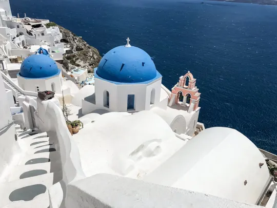 White-washed buildings with blue domes overlook a deep blue Aegean Sea.