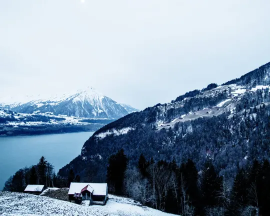 Snow-capped mountains and a lake frame a small cabin in a winter landscape.