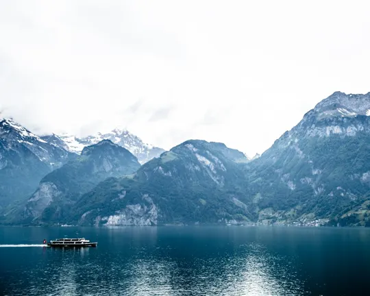 A lone boat glides across a calm lake framed by misty, snow-capped mountains.