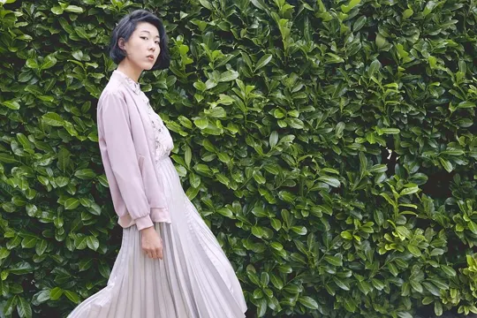 A young woman in a pastel pink jacket and pleated skirt stands against a dense, vibrant green hedge, with soft, diffused light highlighting.