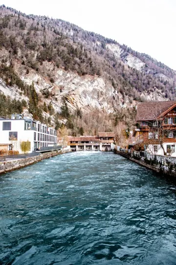 A river flows through a mountain town, framed by stone buildings and forested slopes.