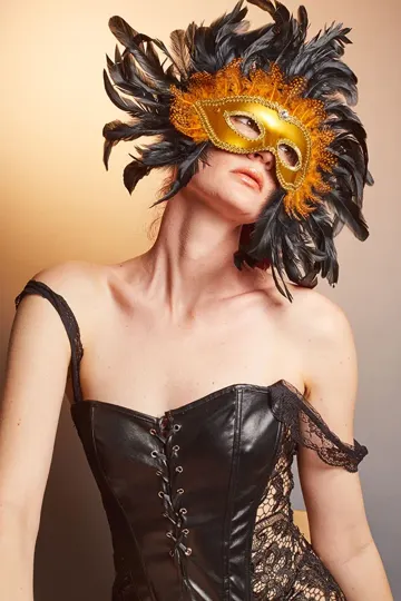 Subject in corset and feathered mask, soft gradient background, warm lighting.