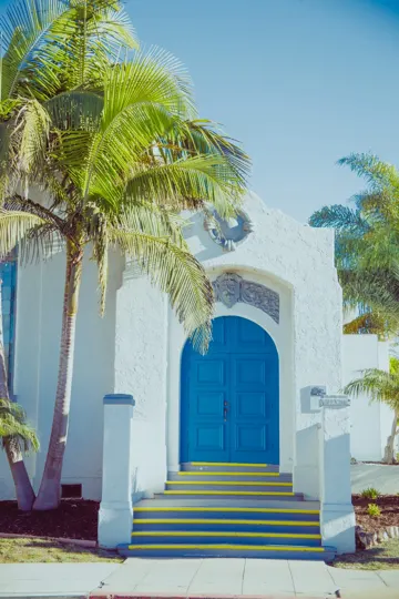 A white stucco building with a blue door, flanked by palm trees under a clear sky.