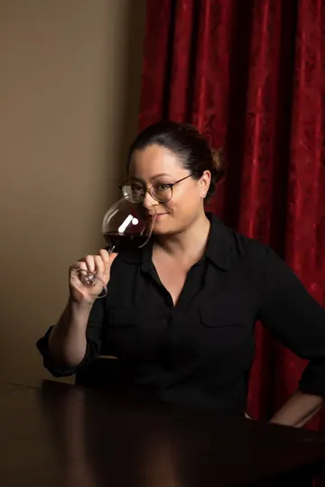 The Cellar Geek in black shirt, holding wine glass, seated at dark table with red curtain backdrop.