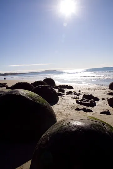 Large, moss-covered boulders frame a sunlit beach with distant hills under a clear sky.