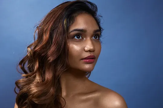 Copper-toned curls cascade against a muted blue backdrop, softly lit.