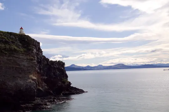 A solitary lighthouse perches on a rugged cliff, overlooking a calm, expansive sea under a partly cloudy sky.