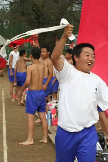 A young man in a white shirt and blue shorts holds a white cloth aloft, surrounded by shirtless men in blue shorts, with red and white.