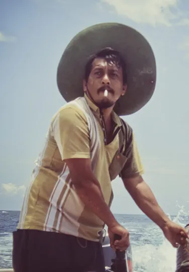 Fisherman steers a small boat under a wide-brimmed hat on a sunlit sea.