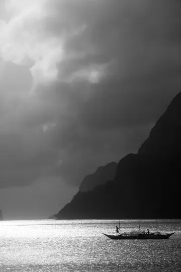 A lone boat glides through a narrow, mountain-framed passage under a moody, overcast sky.
