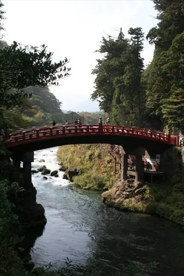 Red wooden bridge spans a rushing river, framed by dense forest under soft daylight.