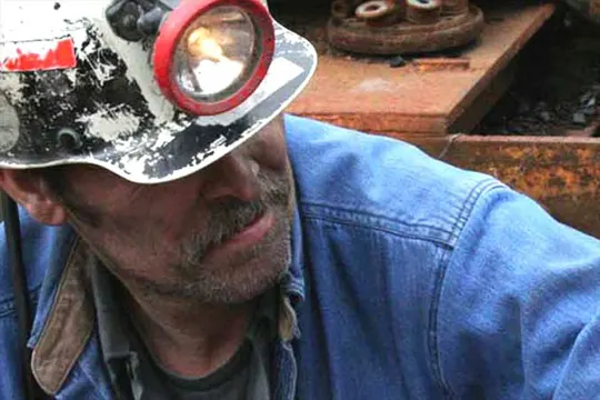 A miner in a blue denim jacket and worn hard hat, illuminated by a headlamp, amidst industrial equipment.