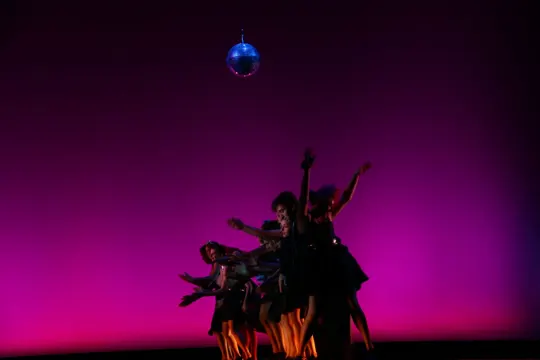 Dancers silhouetted against a vibrant magenta backdrop, illuminated by a suspended disco ball.