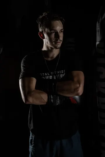 A muscular individual in a "TOTAL FITNESS" shirt stands in dim, dramatic lighting.