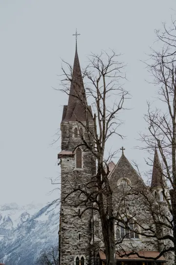 Stone church with tall spire, bare trees, and snow-capped mountains under overcast sky.
