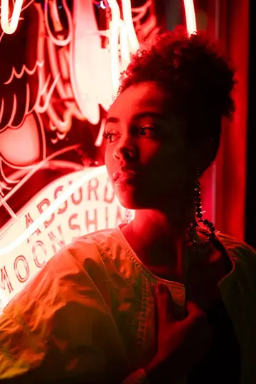 A figure in a white garment stands before a neon-lit sign, bathed in red light.