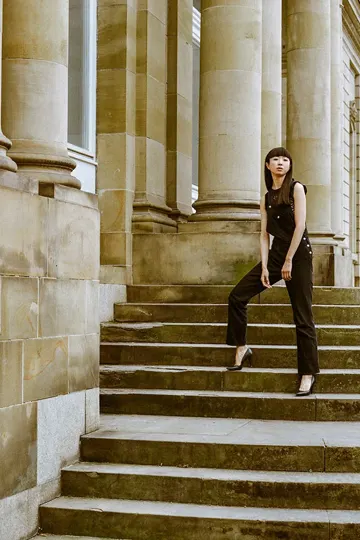 A poised figure in tailored black descends weathered stone steps, soft natural light.