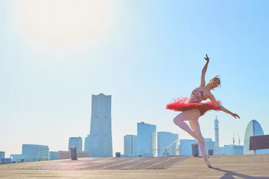 Ballerina in red tutu leaps against sunlit cityscape, shadowed by tall buildings.
