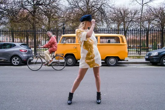 A woman in a gold-embroidered dress and beret stands on a city street, framed by a cyclist and a yellow van.