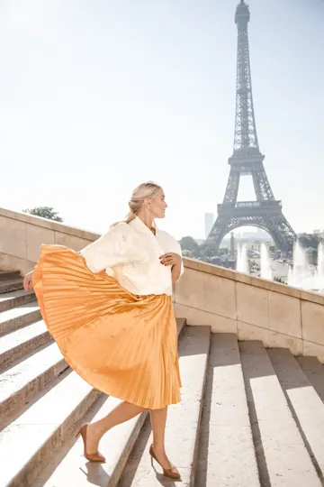 A woman in a white blouse and orange pleated skirt descends steps with the Eiffel Tower and fountains in the background, sunlight casting.