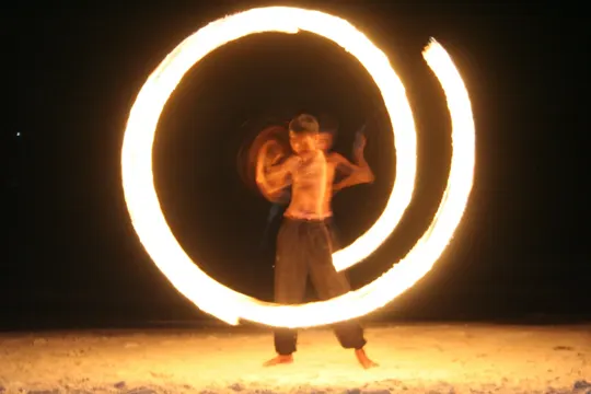 A shirtless performer manipulates a flaming hoop against a dark, sandy backdrop.