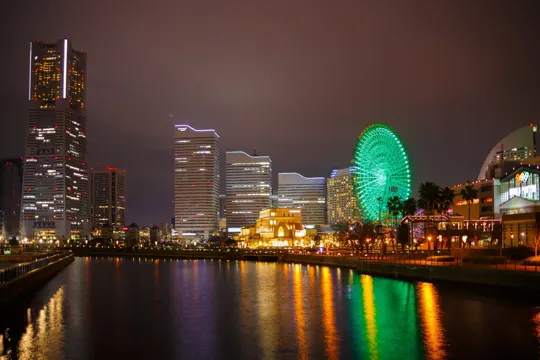 Tokyo skyline at night, illuminated skyscrapers and green Ferris wheel reflected in calm water.