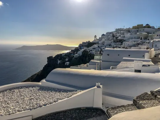 Sunlit white-washed buildings cascade down a cliffside, overlooking the Aegean Sea.
