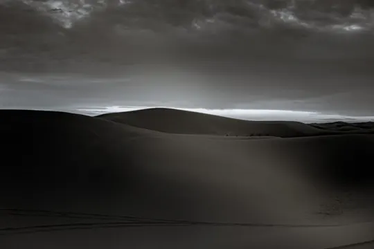 Dunes stretch under a brooding sky, light filters through heavy clouds.
