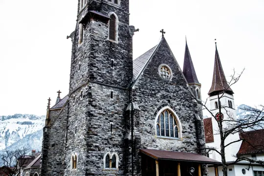 Stone church with tall spires stands against snow-capped mountains under overcast sky.