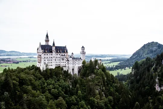 A grand castle perches on a forested hill, framed by distant green valleys under an overcast sky.