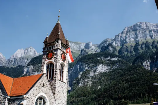Stone church with red roof and Swiss flag, set against rugged alpine peaks under clear blue sky.