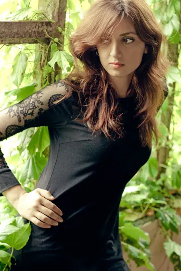 A woman with long, wavy hair stands amidst lush green foliage, illuminated by soft, natural light.